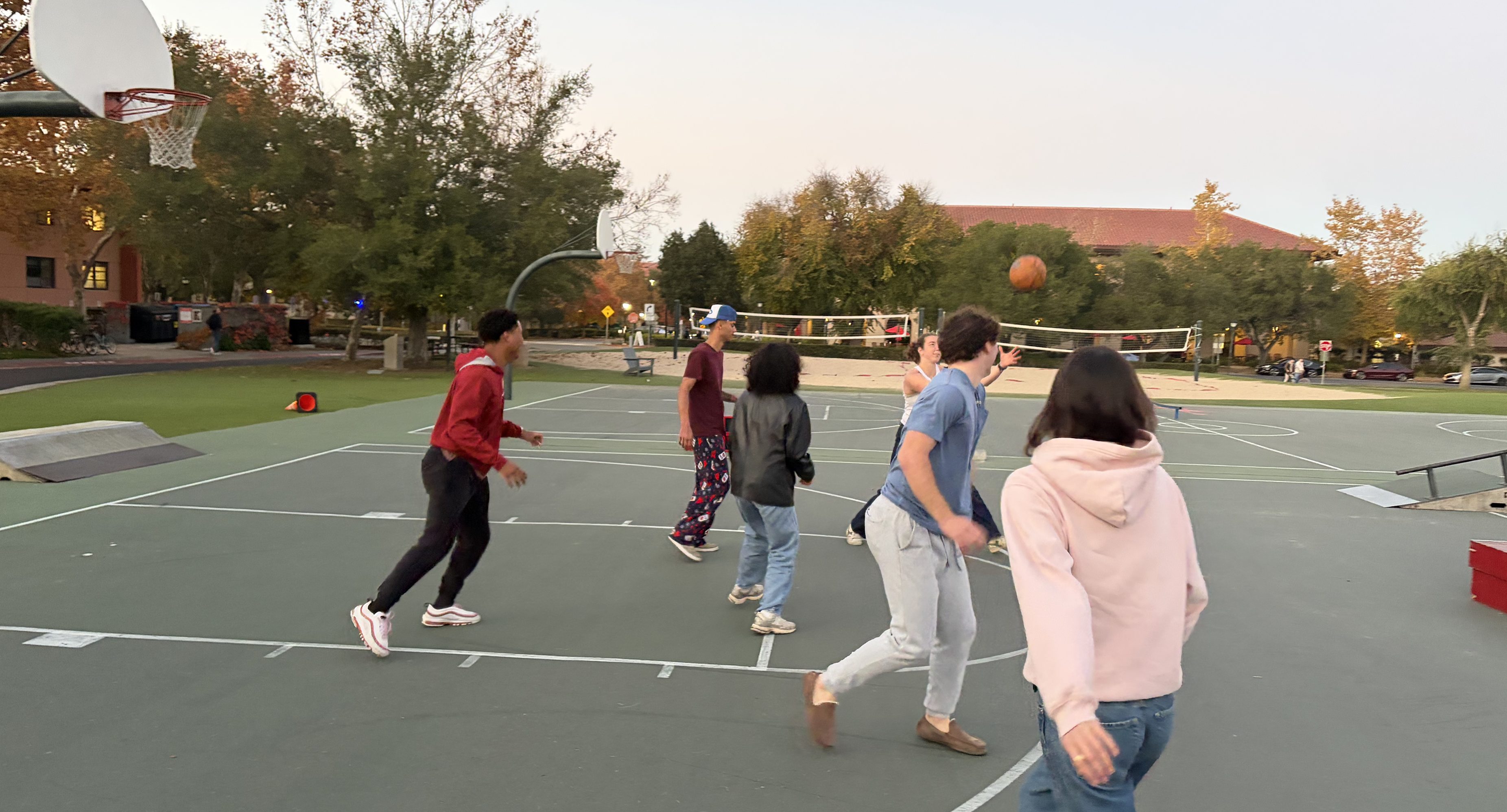 Six students playing basketball on an outdoor court at Stanford during the Sports Day event. A volleyball net and lush trees are visible in the background at dusk.